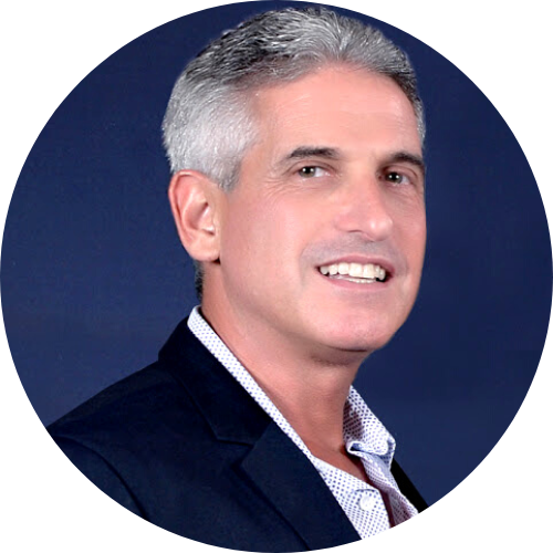 Middle-aged man with short gray hair and a slight smile, wearing a dark jacket over an open-collared shirt, against a dark blue background.