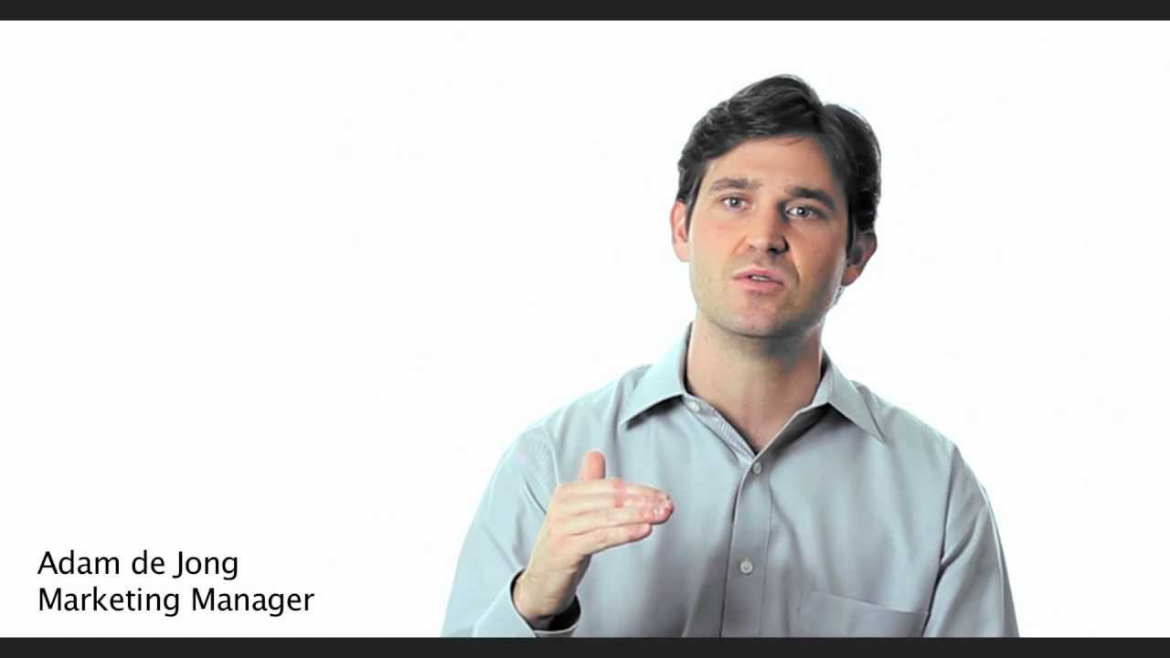 Man in a light blue shirt speaking, with text overlay in the bottom left corner reading "Adam de Jong Marketing Manager." Plain white background.