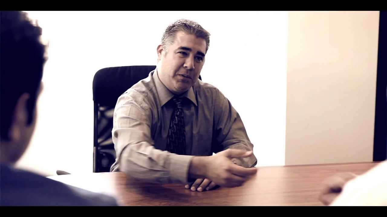 A man in a dress shirt and tie extends his hand across a table toward another person in an office setting.