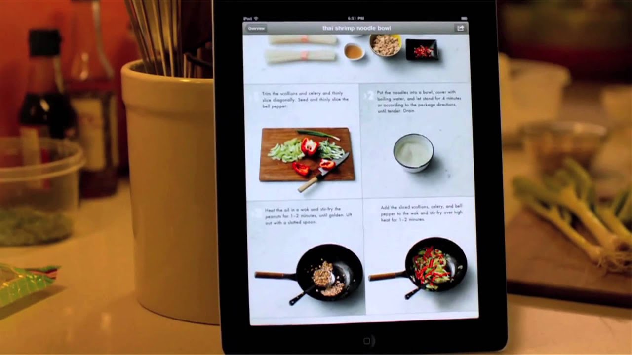 A tablet displaying a recipe for Thai shrimp noodle bowl, featuring images and instructions for preparation steps, positioned on a kitchen counter with various cooking ingredients surrounding it.