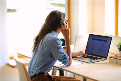 Young woman at desk views laptop screen while holding phone to ear