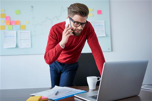 Young bearded man on phoone while looking at laptop screen at work, in red shirt