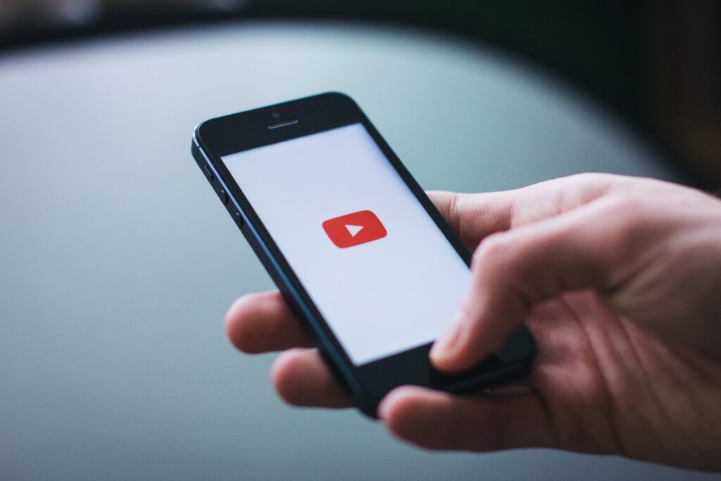 A person holds a smartphone with the YouTube app open, displaying the red play button logo on the screen.