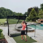 A man, sporting his stylish Nexbelt, practices his golf swing with a net in the backyard, surrounded by a serene pool and lush trees.