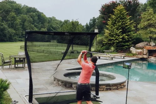 A man, sporting his stylish Nexbelt, practices his golf swing with a net in the backyard, surrounded by a serene pool and lush trees.