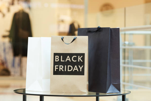 Three shopping bags, including a brown one labeled "Black Friday," sit on a glass table inside a shopping mall, hinting at shoppers’ excitement and hesitation over spending money.