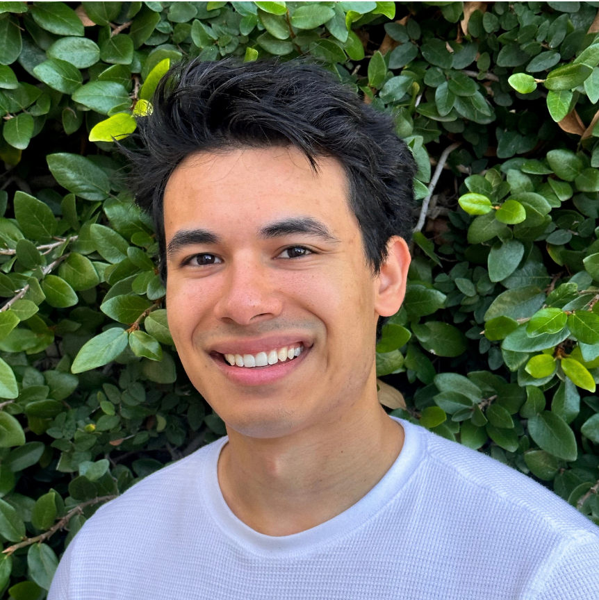 Meet The Team: A person with short dark hair and a white shirt smiles in front of a leafy green background.