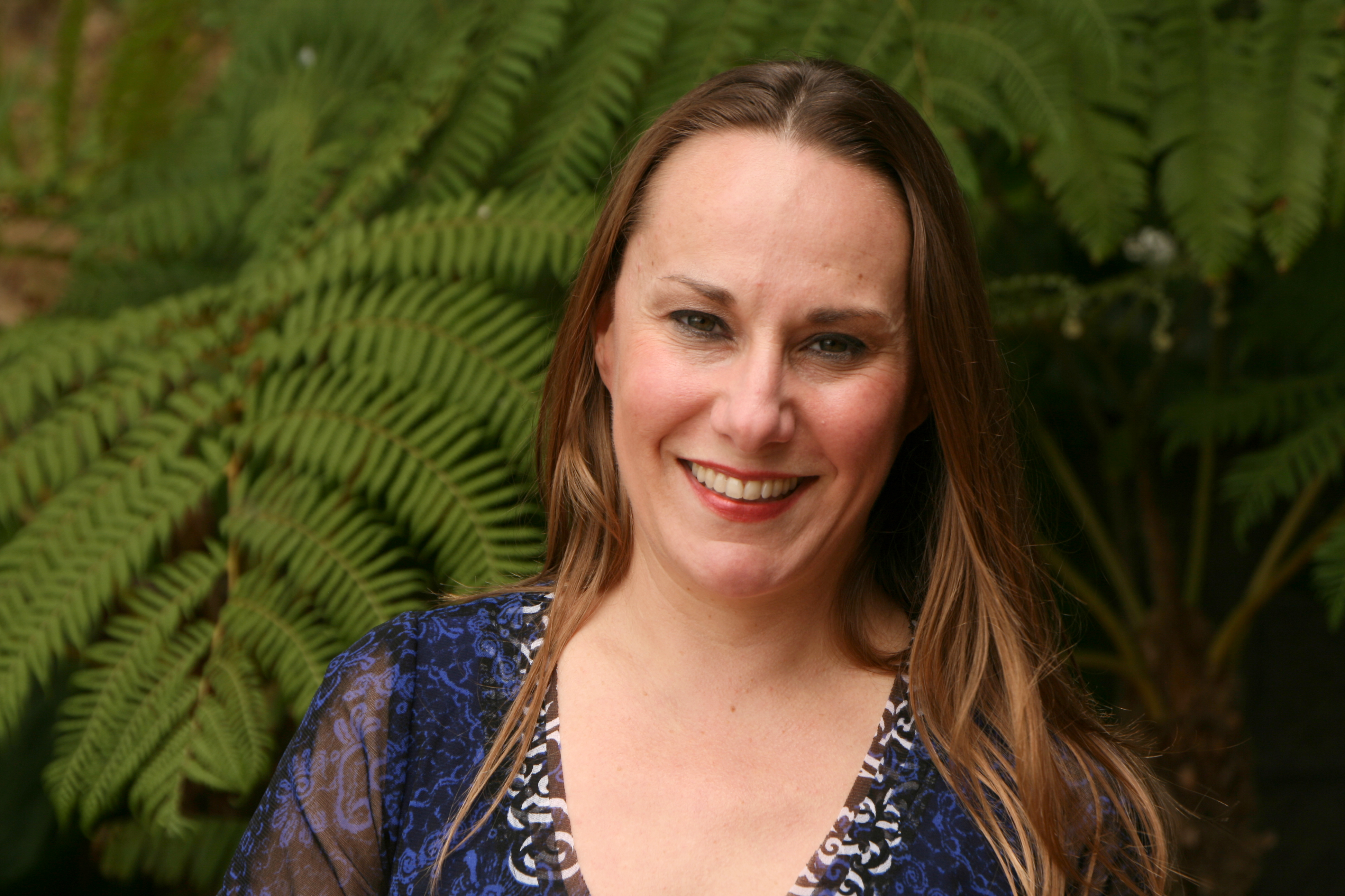 Meet The Team: A woman with long brown hair and a blue patterned shirt smiles at the camera, green ferns in the background.