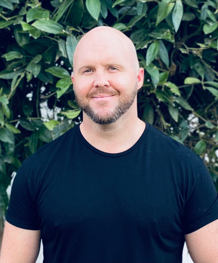 Meet The Team: A man with a shaved head and beard wearing a black t-shirt stands in front of green leafy plants, smiling slightly at the camera.
