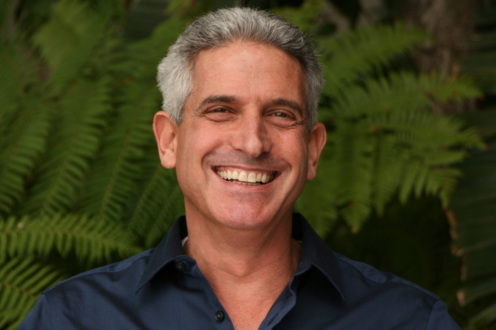 Meet The Team: A middle-aged man with gray hair smiles broadly while wearing a dark blue shirt, standing in front of green leafy plants.