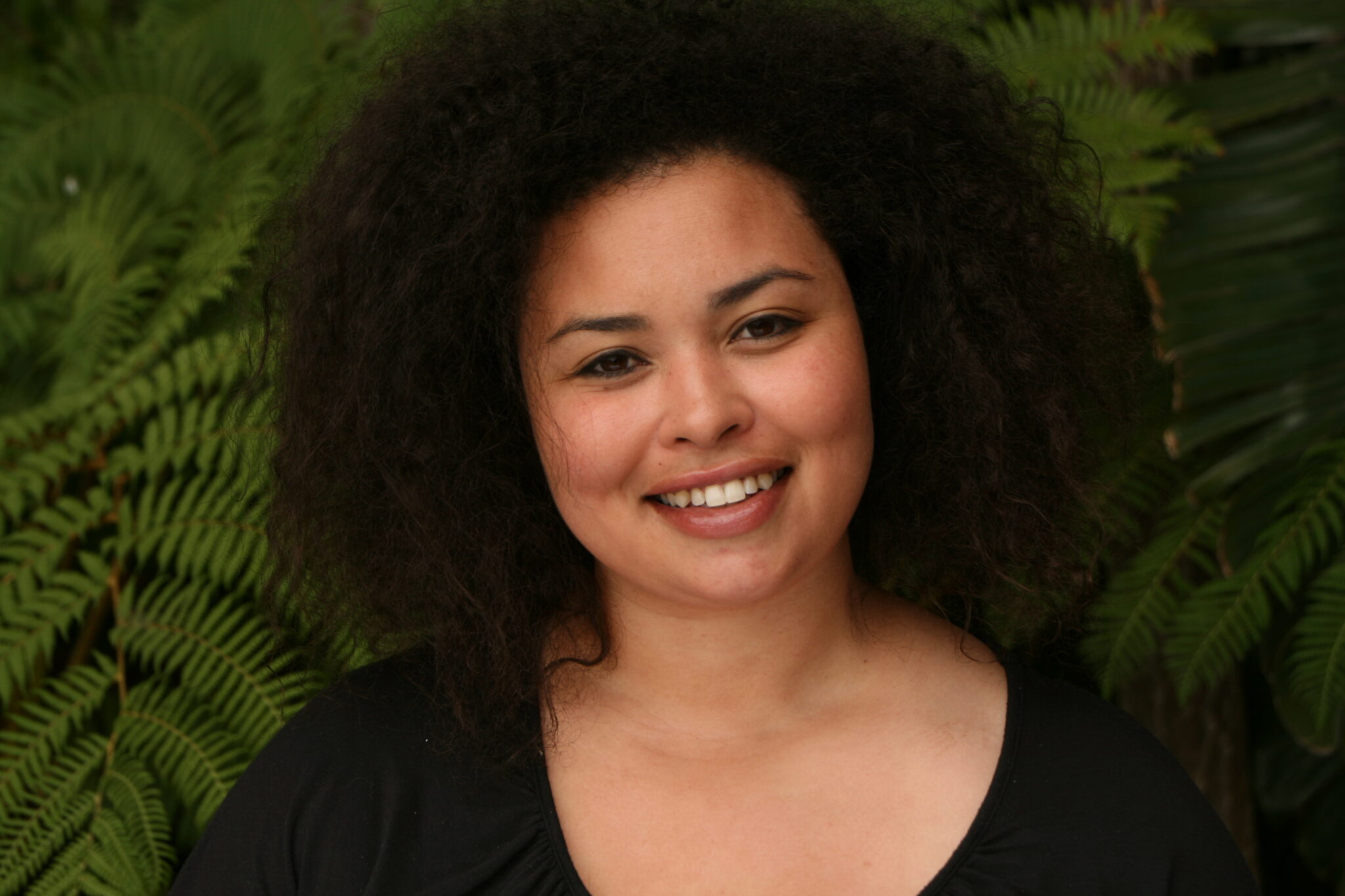 Meet The Team: A person with curly hair and a black top smiles in front of green fern leaves.