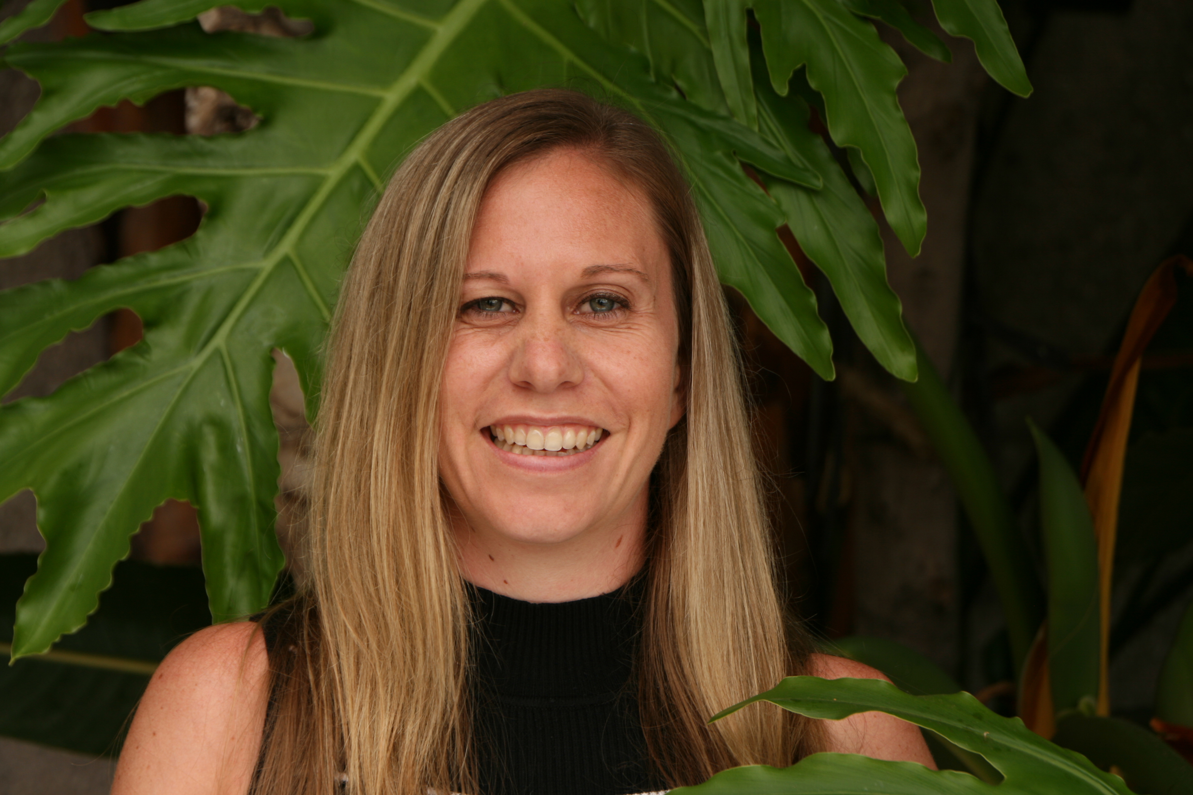 Meet the Team: A woman with long blonde hair and a black sleeveless top smiles in front of large green leaves.