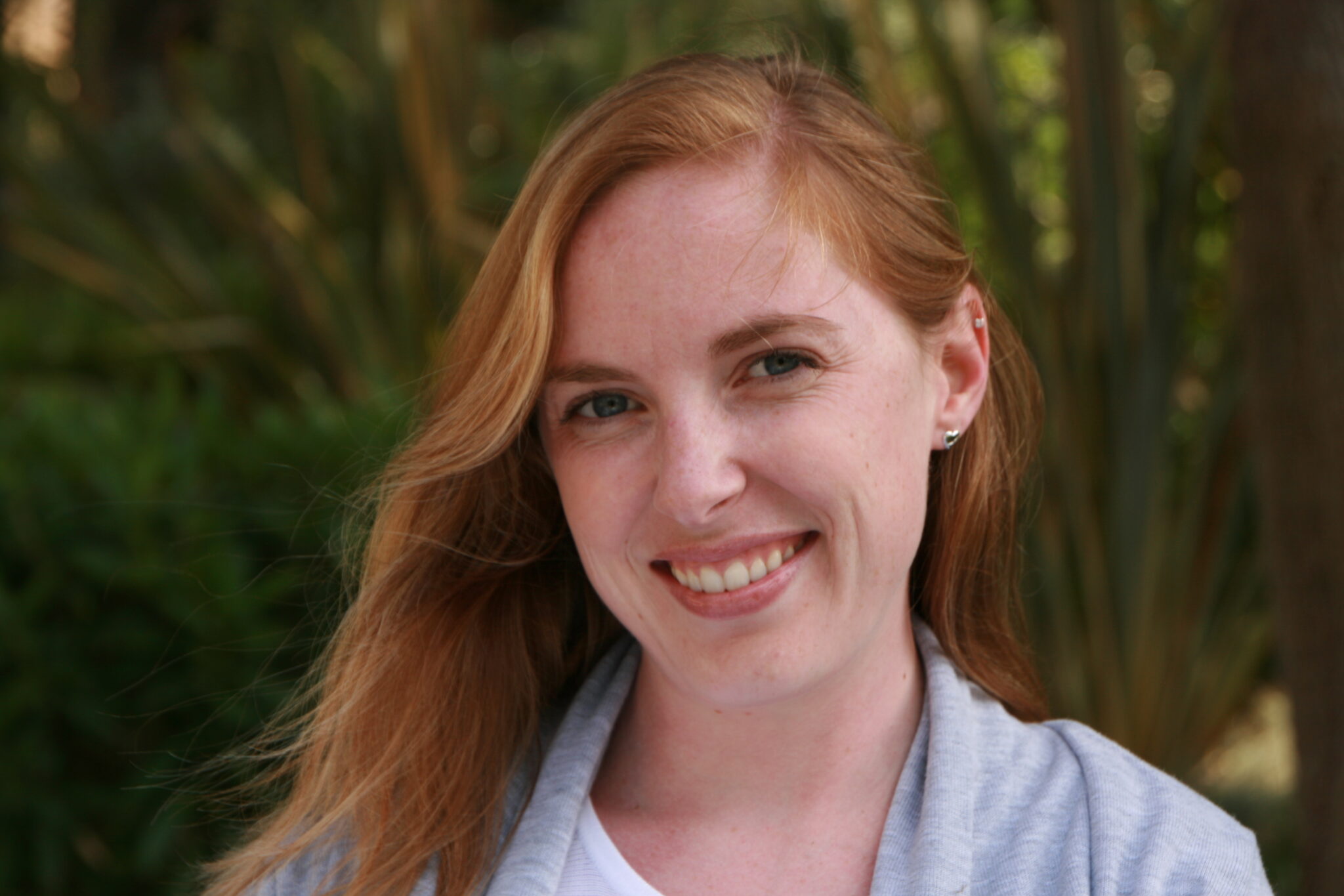Meet The Team: A woman with long red hair smiles at the camera outdoors, standing in front of green foliage. She is wearing a light gray jacket and a white top.