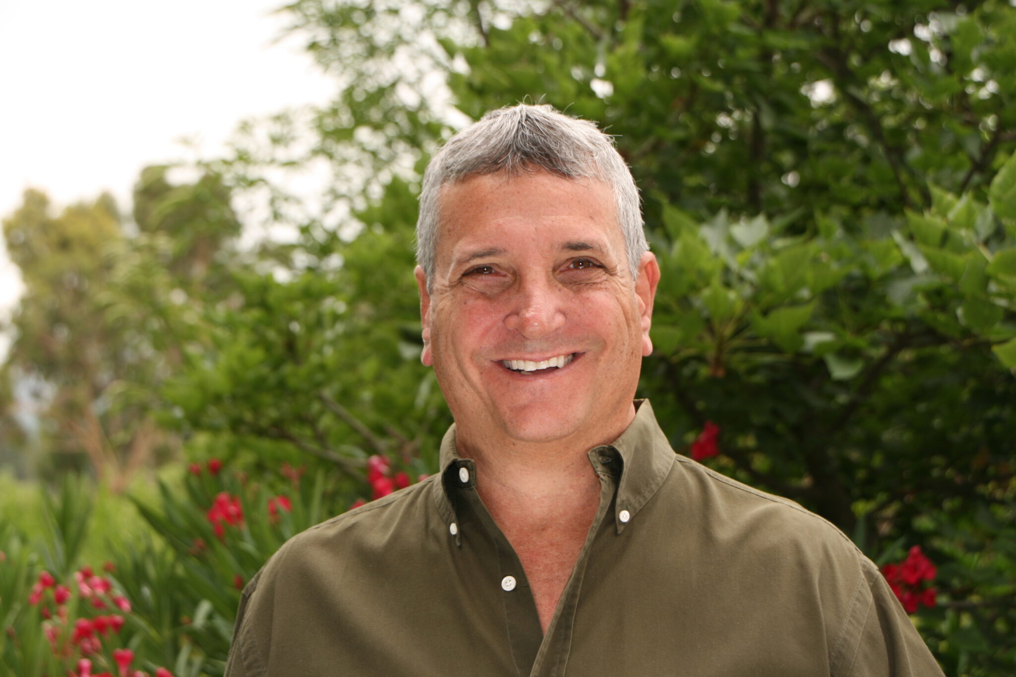 Meet The Team: A man with short gray hair wearing an olive green button-up shirt smiles outdoors in front of green trees and pink flowers.