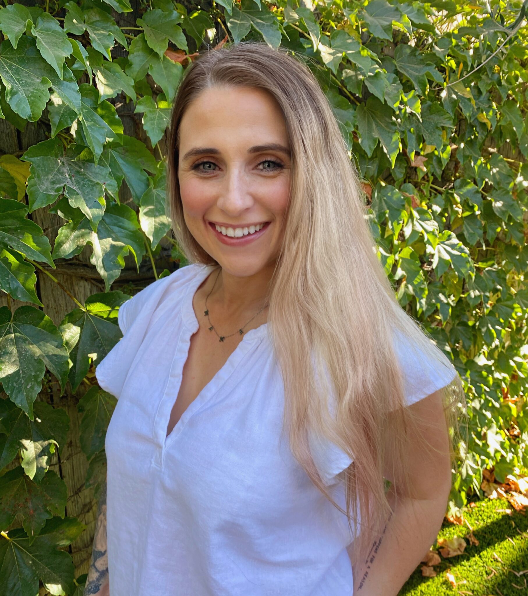 Meet The Team: A woman with long blonde hair wearing a white blouse stands outdoors in front of green leafy foliage, smiling at the camera.