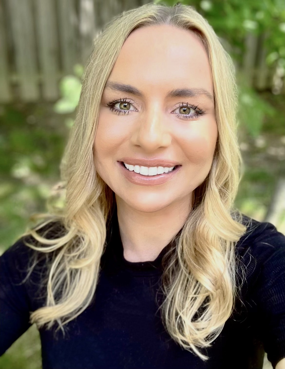 Meet The Team: A woman with long blonde hair wearing a black top is smiling outdoors, with greenery and a wooden fence in the background.