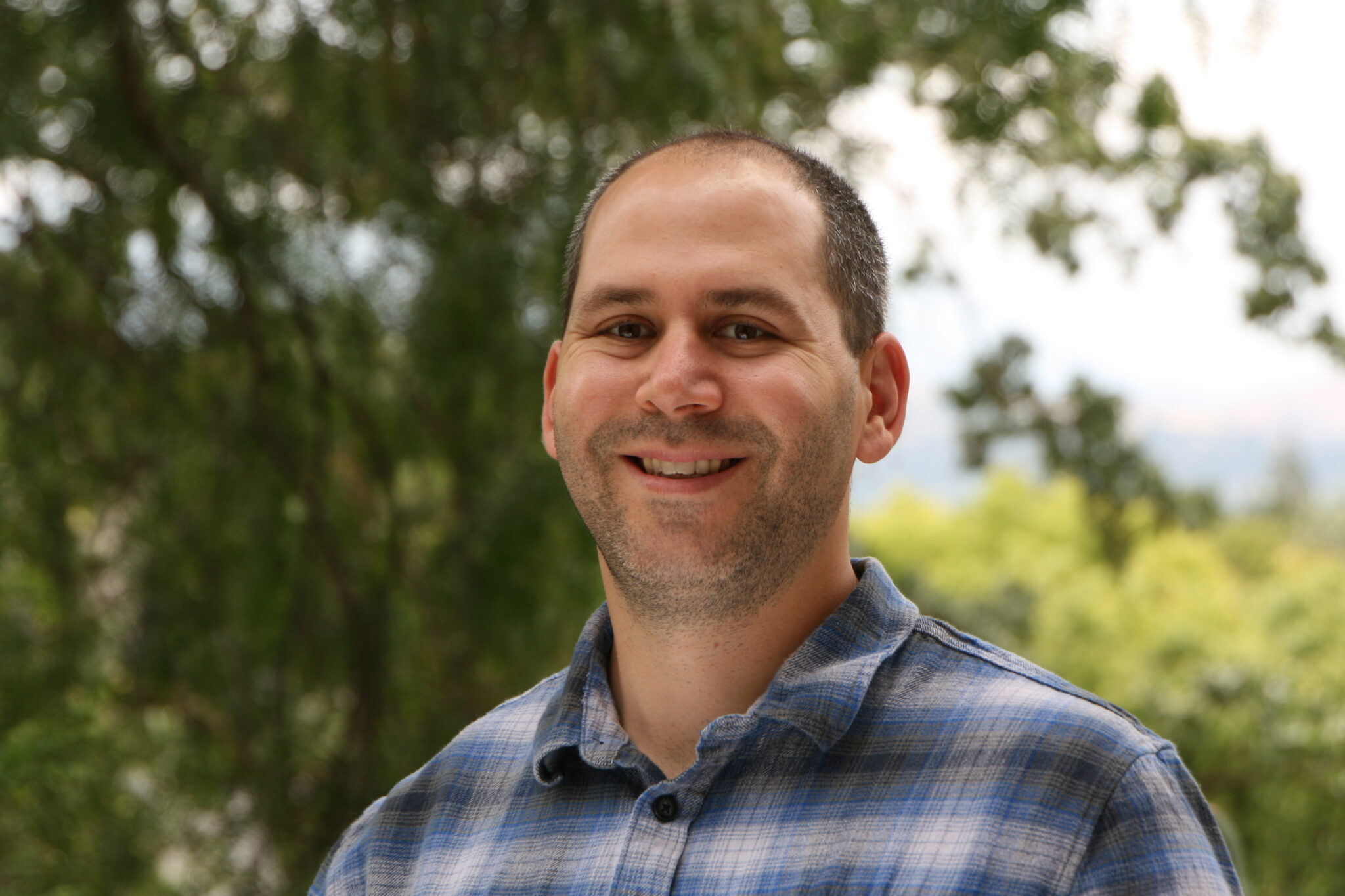 Meet The Team: A man in a blue plaid shirt smiles at the camera outdoors, with green trees and foliage blurred in the background.