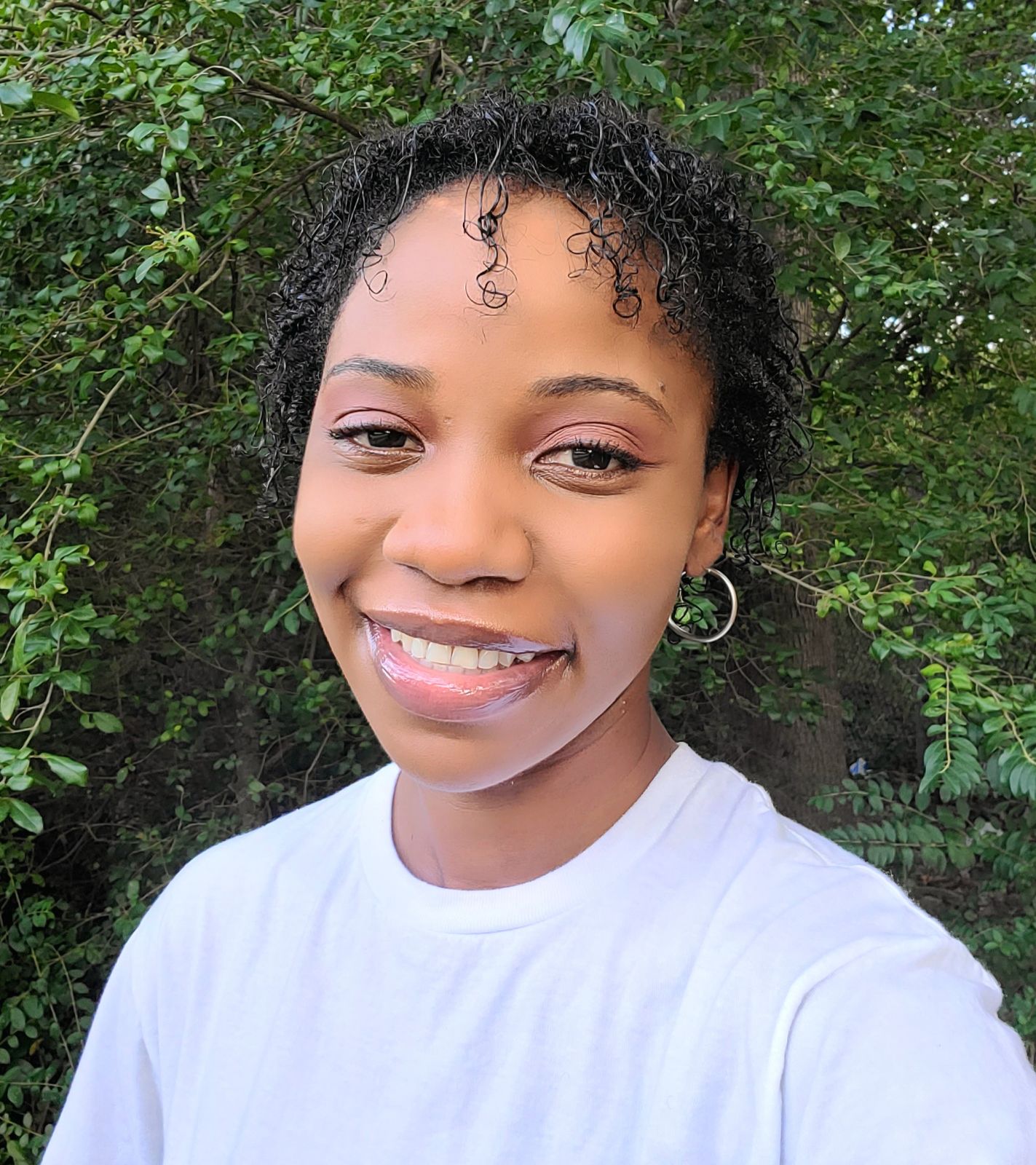 Meet The Team: A woman with short curly hair, wearing a white shirt and hoop earrings, smiles outdoors in front of leafy green trees.