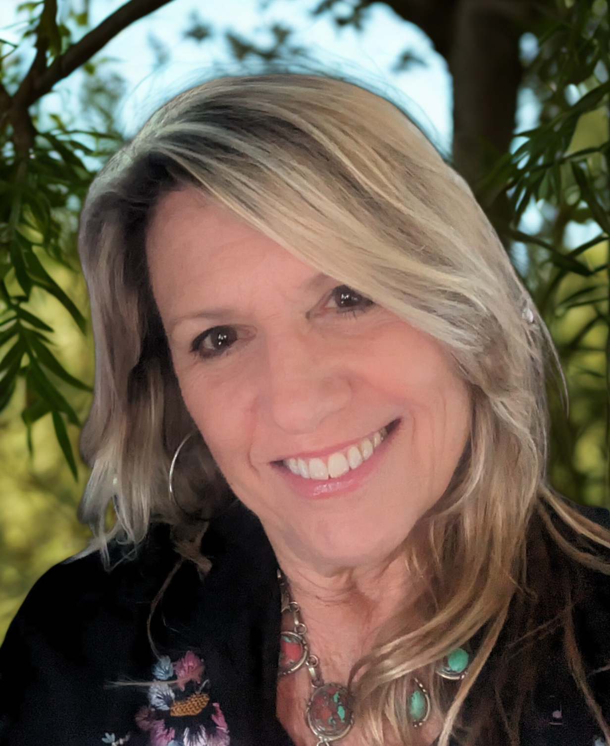 Meet The Team: A woman with long blonde hair, wearing a black floral jacket and turquoise necklace, smiles outdoors with green foliage and a large tree in the background.