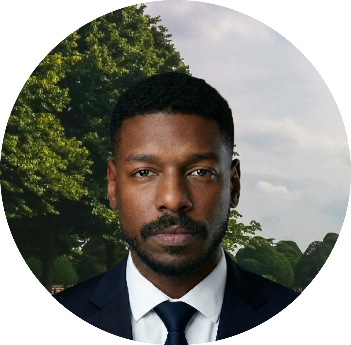 Meet The Team: A man in a suit and tie stands outdoors in front of green trees and a cloudy sky, looking directly at the camera.
