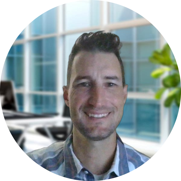 Meet The Team: A man with short brown hair and a plaid shirt smiles at the camera in a modern office, large windows and a plant in the background.