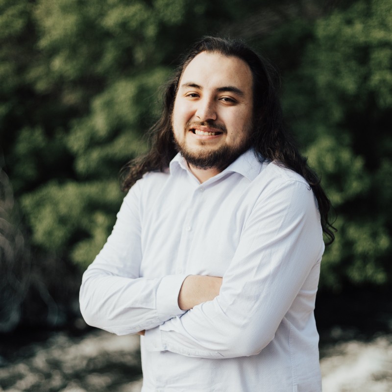 Meet the Team: A person with long dark hair and a beard, wearing a white button-up shirt, stands outdoors with arms crossed, smiling, with greenery in the background.