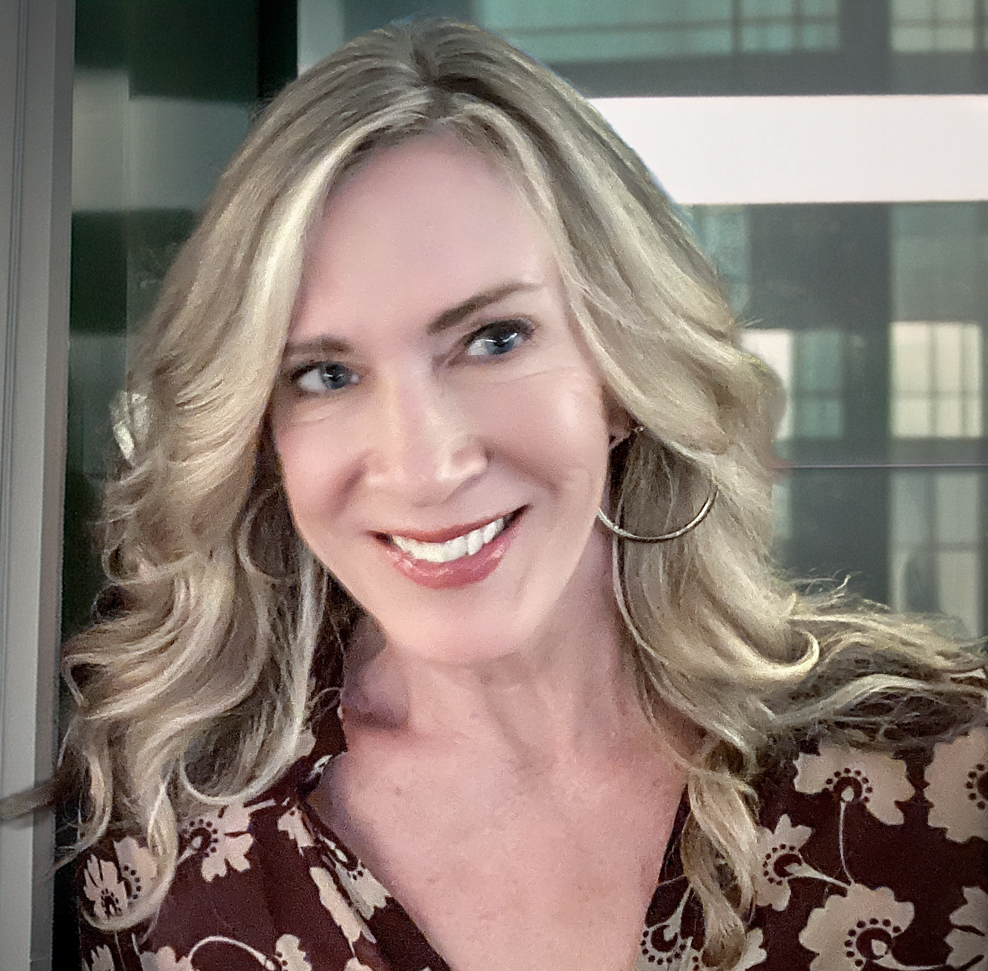 Meet The Team: A woman with long, wavy blonde hair wearing a patterned brown top and large hoop earrings smiles as she stands indoors in front of a window.