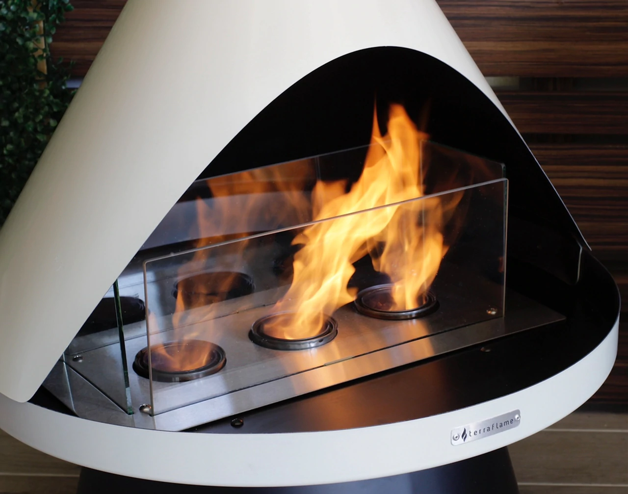 Modern bioethanol fireplace with three circular flame burners inside a white cone-shaped exterior against a dark wooden wall.