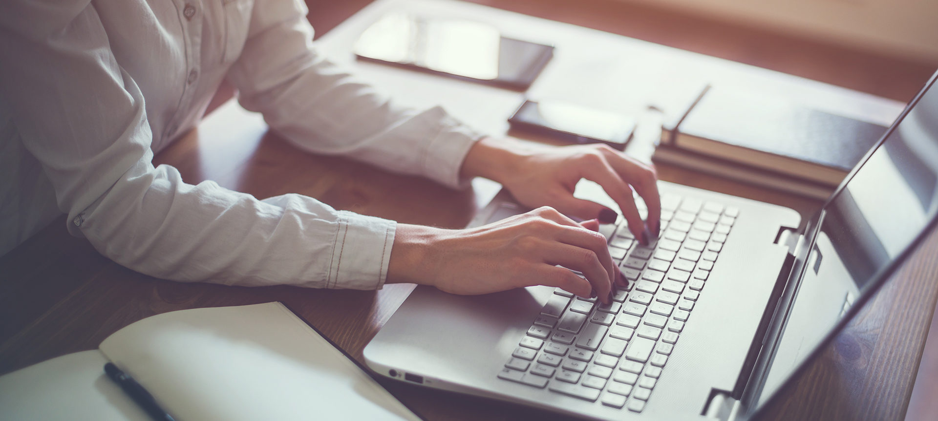 Person in a white shirt typing on a laptop keyboard at a wooden desk with notebooks and materials nearby.